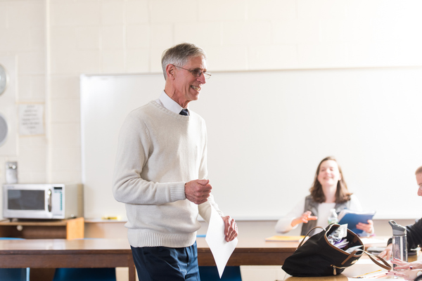Dr. Steven Greenberg smiling and holding a piece of paper while teaching a class with a smiling female student sitting at a desk in background