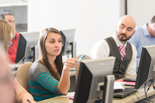a BSU student in management class sits at a desk behind a computer and points a highlighter while speaking. Classmates look on.