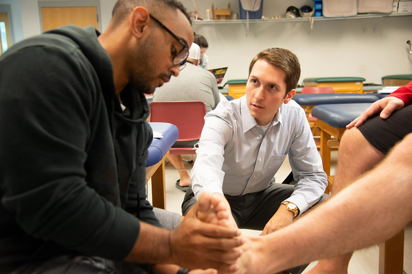 photo of Athletic Training class with 1 student sitting on a medical table, another student holding and looking at the first student's foot and a professor showing the students something on the foot