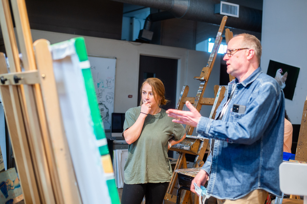 Dr. Collin Asmus and a student looking at and discussing a painting sitting on an easel