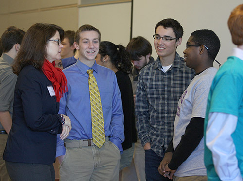 3 high school students talk to a teacher with other students in the background