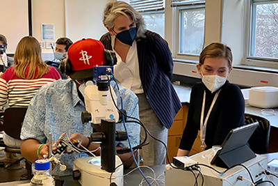 Two students, sitting at a lab table, use a microscope and iPad as their dean looks on.