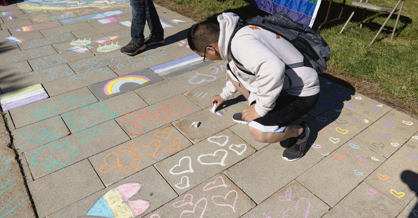 A student draws on the sidewalk with chalk; the sidewalk is decorated with hearts and rainbows for National Coming Out Day