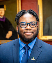 Dhareen Jean-Michel smiling in the BSU Executive Conference Room with short neat dreadlocks, a beard and mustache. He is wearing black rim glasses, a blue suit jacket over a blue button down shirt with a blue tie.