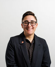 Emily Rose smiling with short dark brown hair and wearing black rimmed glasses and a black blazer over a black print polo shirt