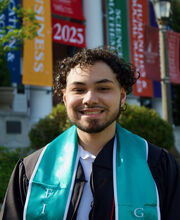 Deontae Jenkins with short curly dark hair and trimmed mustache and beard wearing his BSU graduation gown with his First Generation sash in front of Boyden Hall when it was decorated with Commencement banners for Class of 2025