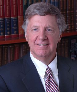 Michael Taylor, UCB President, smiling with short salt and pepper hair wearing a navy blue suit jacket, white button down shirt and a red, white and blue striped tie. There is a full book shelf in background.