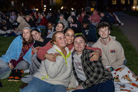 A group of three students hugging among a crowd of others, sitting on the Boyden quad at nighttime