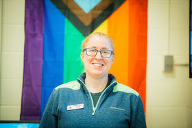Carolyn Taggart, Pride Center Director, standing in front of the pride flag smiling with reddish blonde hair pulled back, wearing blue rimmed eyeglasses and a blue pullover jacket with v-neck zipper
