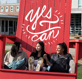 Three students hanging out in the giant red Adirondack chair by Rondileau Student Union. A circle of words above them says, &quot;Yes, I Can.&quot;