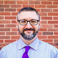 Matt Koskowski smiling with short salt and pepper colored hair, mustache and beard. He is wearing black rim glasses, a blue and white checkered shirt and a purple tie