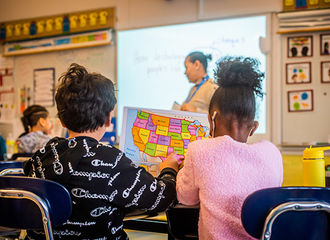 A BSU student teacher stands at front of an Elementary school classroom while 2 students work on a map of the United States