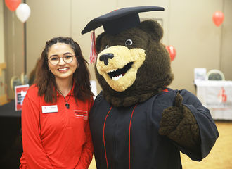 A student smiles next to BSU mascot Bristaco, a bear who is wearing a graduation cap and gown