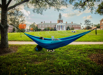 A student hangs in a blue hammock in front of the Boyden Quad — Boyden Hall is in the background