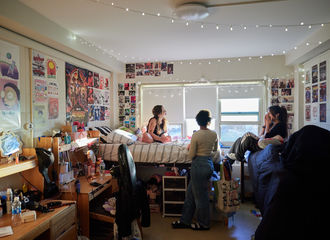 Three students laughing in a dorm room, which is decorated with string lights, photographs, and posters