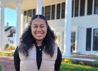 a female student smiles at the camera in front of 161 summer st. She has shoulder-length, curly black hair tied back and is wearing a black shirt with a tan vest over it and a small necklace