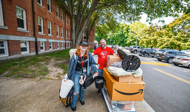 A girl and her father stand smiling while pushing a cart full of items for her dorm room