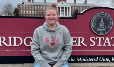 Addy Hodson wears a BSU sweatshirt sitting in front of red Bridgewater State University sign