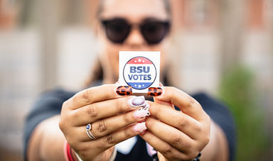 A person is holding a BSU Votes sticker in front of their face with colorful fingernails