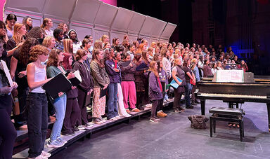 A large group of students sing on a stage next to a piano.