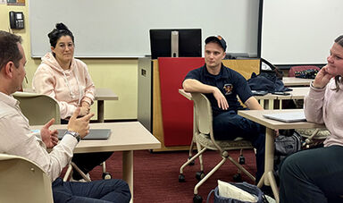 Three students and a professor talk while sitting at tables in a classroom.