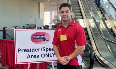 Gabe Elias stands in front of sign wearing a red shirt with hands clasped in front of him