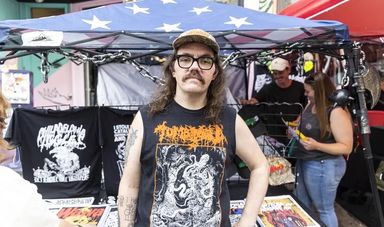 A man with glasses wearing a hat stands in front of a tent selling tshirts