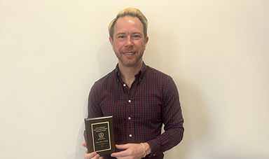 Adam Brieske-Ulenski stands in front of a wall holding his award plaque.