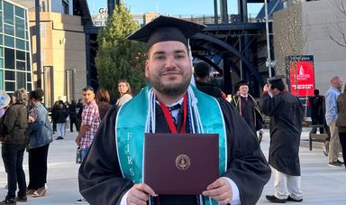 A man wearing graduation cap and gown holds a diploma in front of him