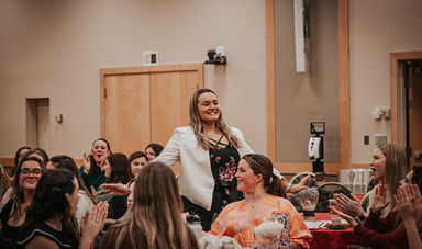 A woman stands smiling holding her hands out while people clap their hands around her