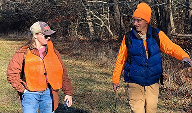 Two people walk through a field next to woods.