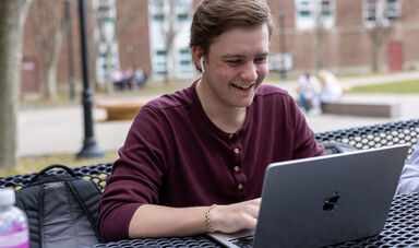 A man wearing a red shirt sits looking at his computer screen and smiles
