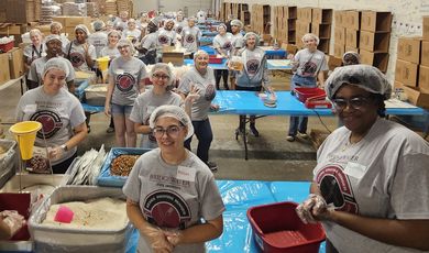 A group of people wearing hair nets stand smiling and packing meals at tables 