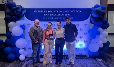 Four people stand in front of a sign smiling 