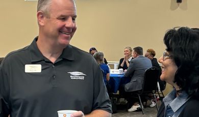 A man stands smiling holding a cup of coffee while talking to a woman who is also miling 