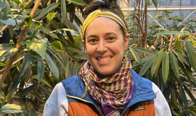 A smiling woman stands in front of plants inside of a greenhouse 