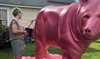 A person stands next to a pink bear and uses a paintbrush to paint 