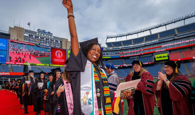 a woman in a cap and gown raises her hand to the sky and smiles 