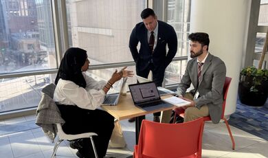 Three people work on a presentation at a table in front of a large window.