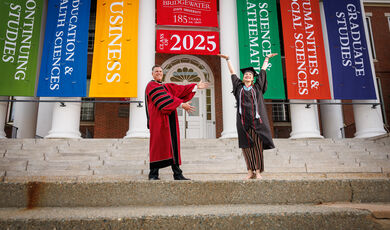 A woman stands on stairs with her hands up in the air, smiling while a man stands next to her also smiling 