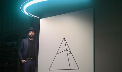 A man stands next to a white canvas with a triangle on it with a blue light above him 