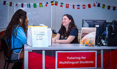 Two women sit at a desk and talk to each other 