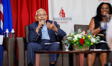 A man sits holding a microphone smiling and talking 
