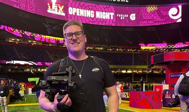 A man stands holding a camera inside of a football stadium 