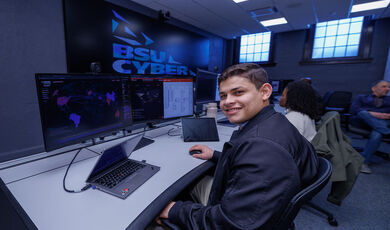 Luis Marin, '26, working at a desk in the BSU Security Operations Center. He's smiling at the camera with short brown hair and wearing a navy blue jacket with khaki pants