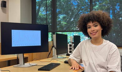 Aaliyah Oliveira smiling sitting at a computer on campus. She has a medium length afro hair style and is wearing a white top.