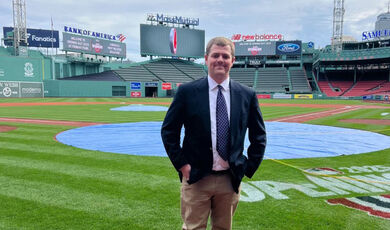 Jesse Frazier, 22, standing on the baseball field at Fenway Park wearing khaki pants, a navy blue blazer over a white button down shirt and a navy blue tie with small print