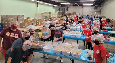 Photo of BSU student volunteers at End Hunger New England packing meals for folks experiencing homelessness. They are spaced out across 4 tables in groups, and are wearing masks, gloves and hairnets and looking at the camera