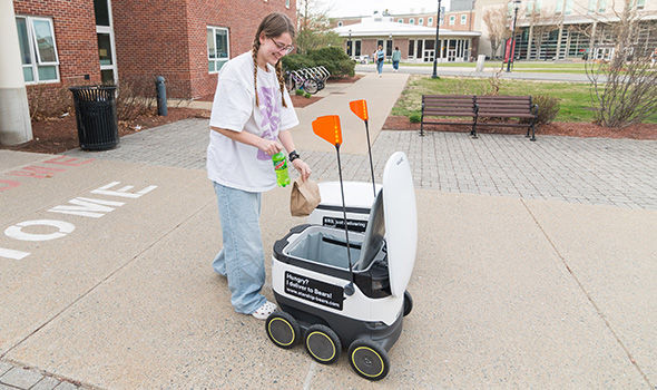 A student takes a bag of food and a soda out of a delivery robot in front of a dorm