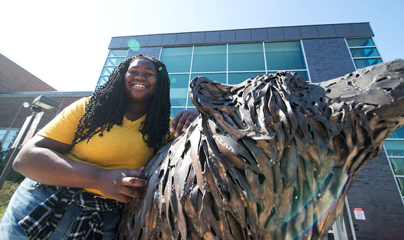 A student smiles brightly while leaning against the bronze bear sculpture in front of the Rondileau Student Union